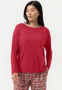 Vrouw in een losse rode lange mouw top en gestreepte broek met rode, beige en witte cirkels, staande met haar handen gedeeltelijk in de zakken.