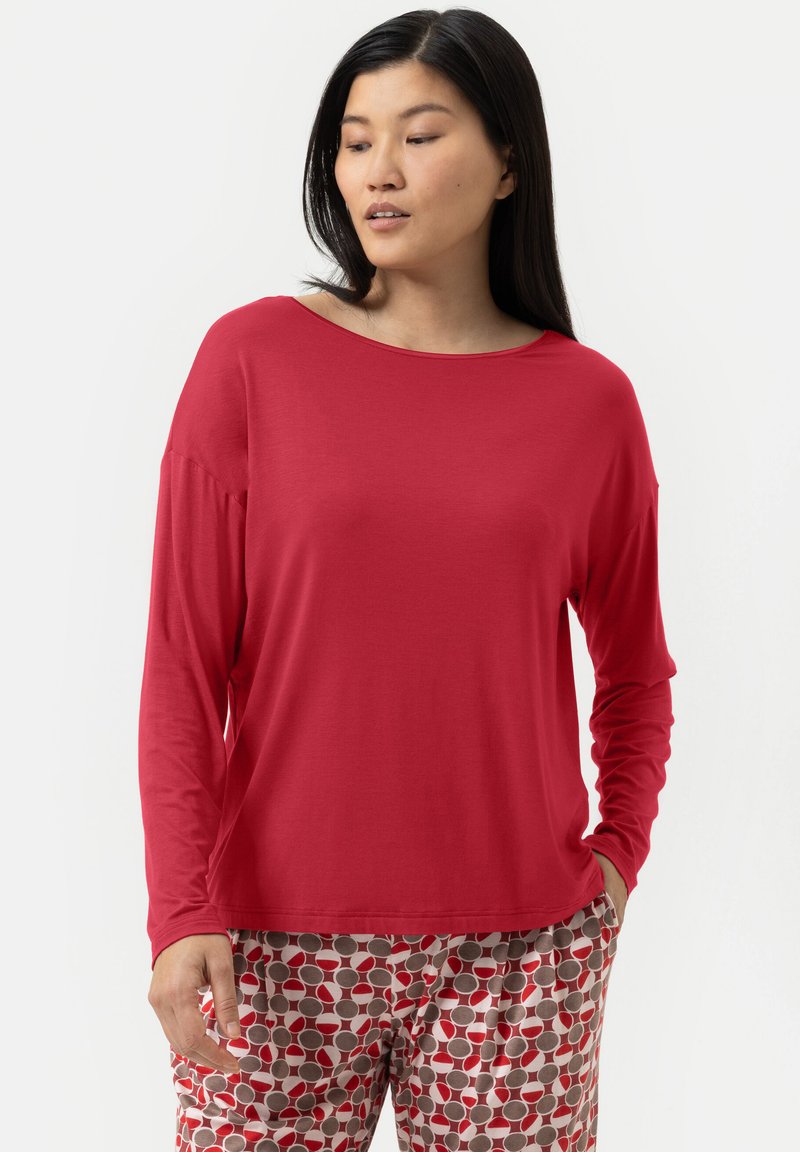 Vrouw in een losse rode lange mouw top en gestreepte broek met rode, beige en witte cirkels, staande met haar handen gedeeltelijk in de zakken.