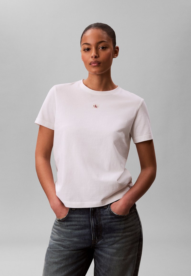 Jeune femme les mains dans les poches, portant un T-shirt blanc uni à manches courtes avec un petit logo rouge sur la poitrine et un jean en denim foncé.