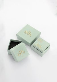 Dva mätovo zelené boxy so zlatým brandingom "elli", jeden otvorený, ukazujúci čiernu vnútornú výplň. Väčší box je štvorcový, menší je obdĺžnikový.