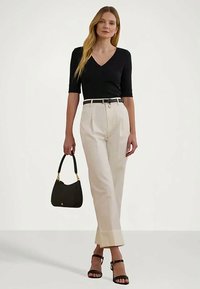 Fekete V-kivágású felsőt viselő nő, bézs magas derekú nadrágban, fekete övvel, fekete magassarkú szandálban, és fekete kézitáskával a kezében.