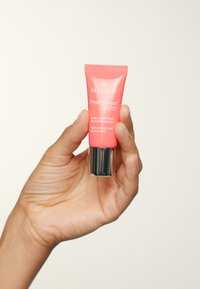 Tube rose de gel de baume pour les yeux NUXE Prodigieuse Boost avec un fond argenté, présentant une texture lisse et un texte imprimé, tenu dans une main contre un fond clair.