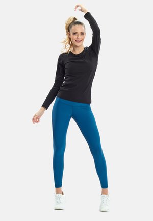 Zwarte lange mouwen top en blauwe leggings. Top heeft een ronde halslijn; leggings zijn strak en glad. Model draagt witte sneakers.