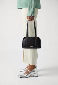 Borsa in pelle nera con doppi manici superiori e dettagli dorati, abbinata a pantaloni aderenti di colore chiaro e scarpe con tacco quadrato argentato lucido.