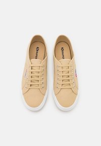Beżowe trampki z płótna o niskim kroju, z metalowymi oczkami na sznurówki oraz białymi gumowymi podeszwami. Logo widoczne na wkładce i boku.