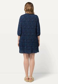 Tummansininen mekko, jossa on rypytetty yläosa, kolmen neljäsosan hihat ja levenevä helma. Sisältää monivärisen täpläkuvion kauttaaltaan.