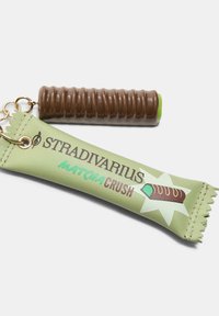Kľúčenka s hnedým kúskom vo forme maškrty so zeleným koncom, pripevnená na zelený kožený štítok s nápisom "Stradivarius Matcha Crush" vytlačeným čiernou farbou.