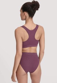 Vrouw met krullend haar in een knot, gekleed in een bijpassende paarse ribbel sportbeha met racerback en een set van hoge taille ondergoed, met de rug naar de kijker gekeerd.