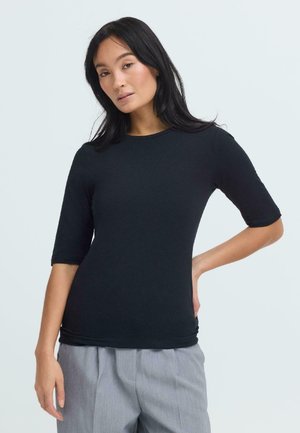 Vrouw met lang donker haar, gekleed in een zwarte nauwsluitende shirt met halflange mouwen en lichtgrijze broek, poseert met één hand op haar heup.