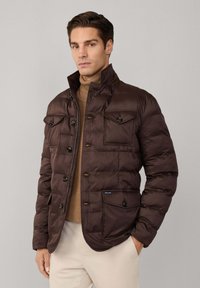 Chaqueta acolchada marrón con cuello alto, dos bolsillos en el pecho con botones y cierre frontal con cremallera. La tela tiene una textura suave y un brillo sutil.