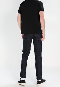 T-shirt noir et jeans bleu foncé, coupe ajustée. Les jeans disposent de poches arrière avec des détails de couture. Des baskets noires décontractées complètent le look.