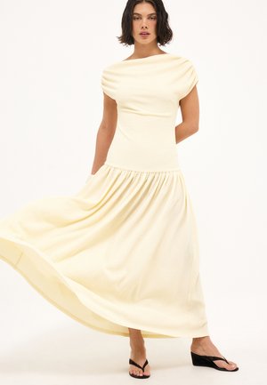 Kobieta w kremowej, teksturowanej sukience midi z marszczoną talią, luźną spódnicą i czarnymi sandałami na koturnie, stojąca na białym tle.
