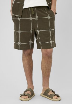 Homme portant un short vert olive avec un motif quadrillé blanc brodé, un gilet assorti et des sandales vert olive à boucle double.