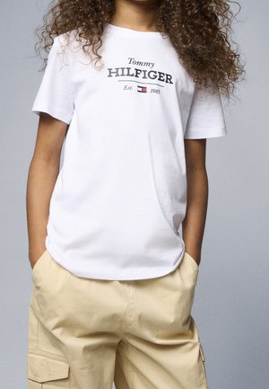 Person med krøllet hår bærer en hvid Tommy Hilfiger t-shirt og beige cargobukser med hænderne i lommerne mod en grå baggrund.