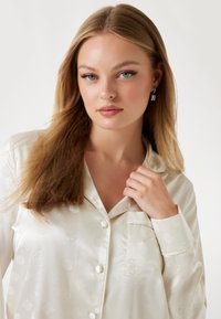 Weißes Satin-Pajama-Oberteil mit einem dezenten Blumenmuster, langen Ärmeln, Knopfverschluss und einer Brusttasche mit Paspelierungen.