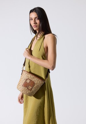 Kobieta w oliwkowej, bezrękawnikowej sukience trzymająca plecioną torbę na ramię w stylu kosza z elementami ze skóry na jednolitym tle.