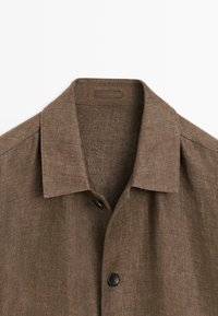 Chemise en tissu chevrons marron avec col pointu et bouton noir visible, gros plan sur la partie supérieure avant.