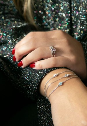 Bagues en argent et un bracelet avec trois accents de pierres ; teint de peau visible, ongles rouges, sur un fond pailleté sombre.