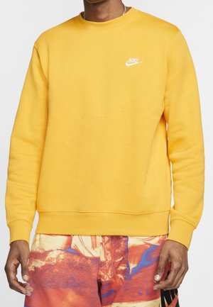 Homme portant un sweat-shirt Nike jaune vif à manches longues et un pantalon à motifs rouges, bleus et beiges, sur un fond uni.