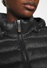 Czarna pikowana kurtka z wysokim kołnierzem, błyszczącą fakturą, suwakiem z przodu i pomarańczowym akcentem logo na ściągaczu suwaka.