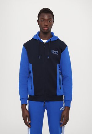 Felpa con zip color navy e blu con logo frontale, tasche e cappuccio con cordino, abbinata a pantaloni del tuta blu con strisce laterali.