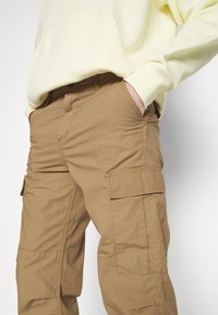 Personne portant un sweat-shirt jaune clair et un pantalon cargo marron avec de grandes poches latérales, une main dans la poche, vue de la taille à la cuisse.