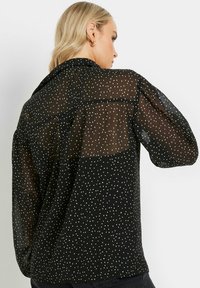 Zwarte doorschijnende blouse met witte stippen, lange pofmouwen en een hoge halslijn; heeft een solide paneel op de bovenrug voor contrast.