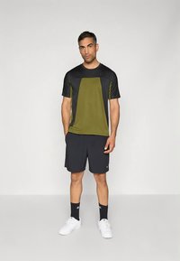 Camisola desportiva de mangas curtas verde e preta com design de painéis contrastantes, combinada com calções pretos e ténis brancos. Meias escuras completam o visual.