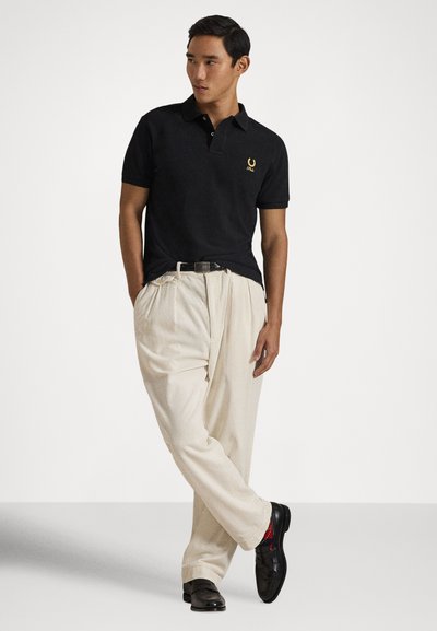 Vyras juoda polo marškinėliais su mažymu logotipu, dėvintis smėlio spalvos plisuotas kelnes, juodą diržą, juodus batus ir raudonas kojines, stovintis prieš baltą foną.