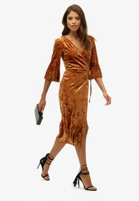 Robe portefeuille en velours orange brûlé avec des manches évasées trois-quarts, taille cintrée et fente latérale, assortie à des sandales à talons noires à lanières.