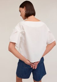 Blusa di cotone bianca con maniche corte e gonfie legate ai gomiti, caratterizzata da un ampio scollo e una vestibilità rilassata. Abbinata a pantaloni corti in denim.