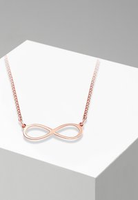 Collana in oro rosa con pendente a simbolo dell'infinito su una delicata catena, esibita su un supporto geometrico bianco.