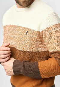 Pull en tricot présentant des rayures horizontales crème, beige et marron. Tissu texturé avec des poignets côtelés et un petit logo sur la poitrine.