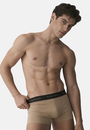 Giovane uomo con corpo tonico che indossa boxer beige con fascia nera con la scritta "Vincent Creation", che sistema la fascia con una mano.