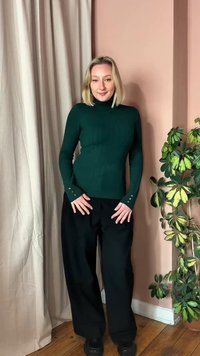 Svetlolasa ženska, oblečena v temno zelen pulover z visokim ovratnikom in črne široke hlače, stoji v sobi z roza stenami, lesenim tlemi in lončnico.