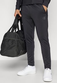 Schwarze Sporthose mit schmalem Schnitt, aus glattem Stoff gefertigt. Verfügt über seitliche Taschen und ein dezentes Logo. Kombiniert mit einer schwarzen Sporttasche und weißen Sneakern.