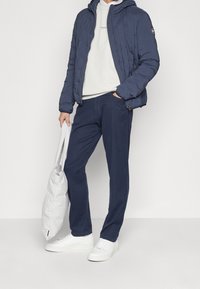 Persoon draagt een marineblauwe pufferjas, licht sweatvest, marineblauwe broek, witte sneakers, houdt een lichtgrijze gewatteerde tas in één hand vast, andere hand in de zak.