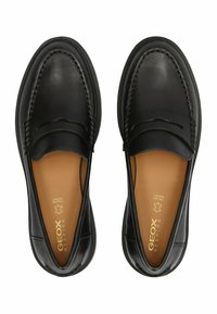 Mocasines de cuero negro con una textura suave, que presentan detalles de costura y un logo dorado en una plantilla color beige, manteniendo un diseño clásico.