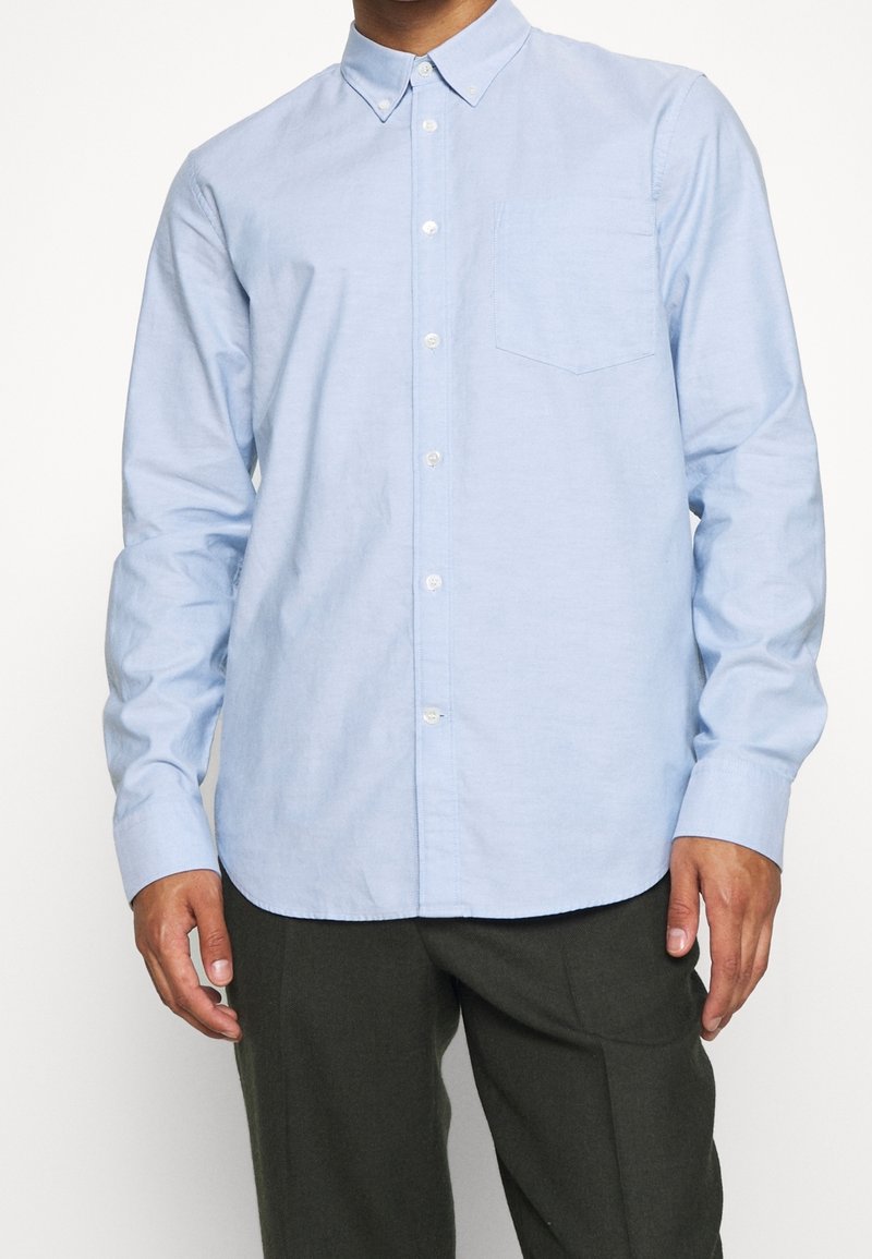 Homme portant une chemise bleu clair à boutons avec poche poitrine et un pantalon vert foncé, debout devant un fond blanc.