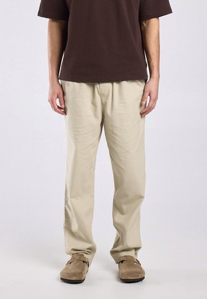 Man draagt beige loszittende broek, bruin kortemouwen overhemd en lichtbruine suède instapschoenen, staat voor een effen achtergrond.