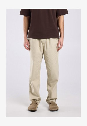 Man draagt beige loszittende broek, bruin kortemouwen overhemd en lichtbruine suède instapschoenen, staat voor een effen achtergrond.
