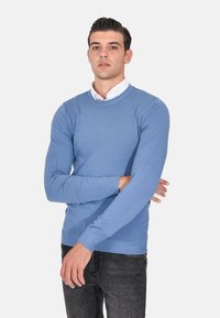 Maglione azzurro chiaro con scollo a giro e maniche lunghe. Realizzato in materiale morbido, presenta polsini a coste e una vestibilità aderente. Indossato sopra una camicia bianca.
