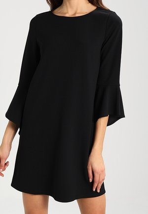 Femme portant une robe noire mi-longue avec des manches cloches 3/4 et un col rond, sur un fond clair uni.