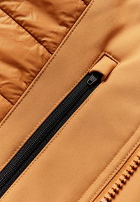 Chaqueta acolchada de color naranja, con un acabado texturizado, un zipper negro y un material suave en el panel al lado del zipper.