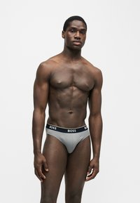 Homme musclé debout sur fond uni, portant un slip gris BOSS avec une ceinture noire, légèrement tourné vers la gauche avec une expression détendue.