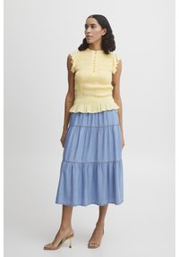 Top amarillo con volantes en las mangas y detalle de botones, combinado con una falda de mezclilla azul clara con volantes. Sandalias de tacón beige completan el conjunto.