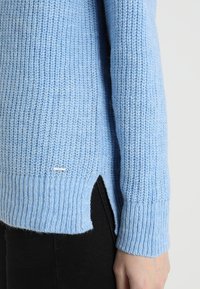 Maglione lavorato a maglia azzurro chiaro con texture a coste, fessure laterali e un piccolo dettaglio in metallo sul fondo. Design morbido e casual.