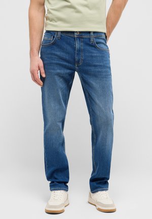 Mann trägt blaue Straight-Leg-Jeans, weiße Sneakers und ein hellbeiges T-Shirt vor einem schlichten hellen Hintergrund.