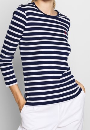 Kobieta ubrana w granatowo-biały, poziomo prążkowany top z długim rękawem z czerwonym haftowanym logo, w połączeniu z białymi spodniami, z rękami w kieszeniach.