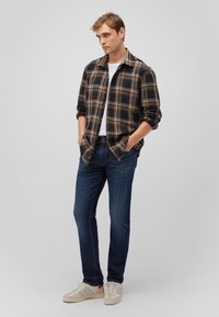 Schwarzes und goldenes karierte Flanellhemden, weißes Rundhals-T-Shirt, blaue Jeans und beige Sneaker mit blauen und roten Akzenten.